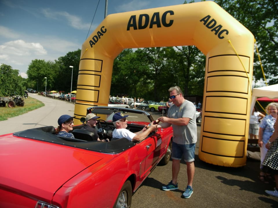 Unser Vorstand begrüßt Gäste in einem roten Muscle Car zu unserem Oldtimertreffen unter dem ADAC-Torbogen.